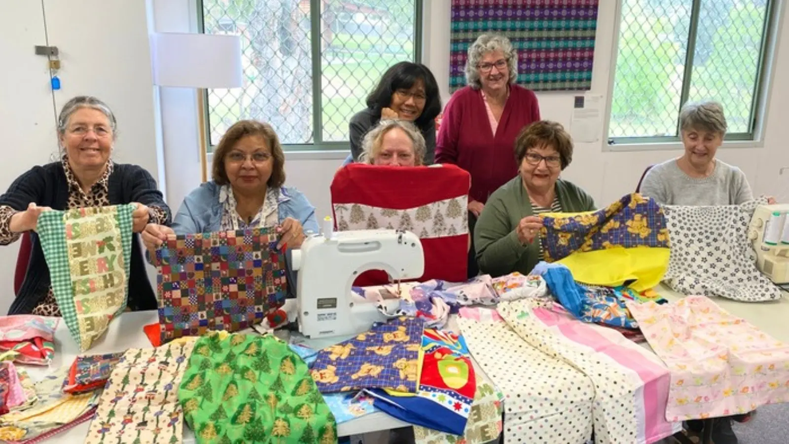 Seven women at a table covered in bright fabric offcuts, holding up patchwork bags they have made, with a sewing machine in the middle