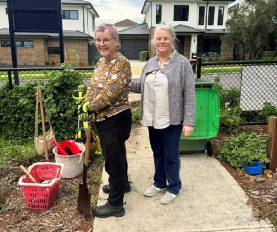 Two volunteers standing on the garden path beside a green bin, with buckets and tools at their feet