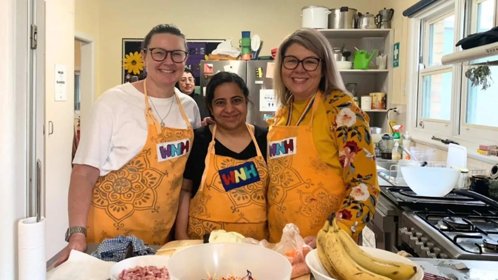 Three volunteers in yellow Watsonia Neighbourhood House aprons standing together in the kitchen behind bowls of ham, salad and bananas