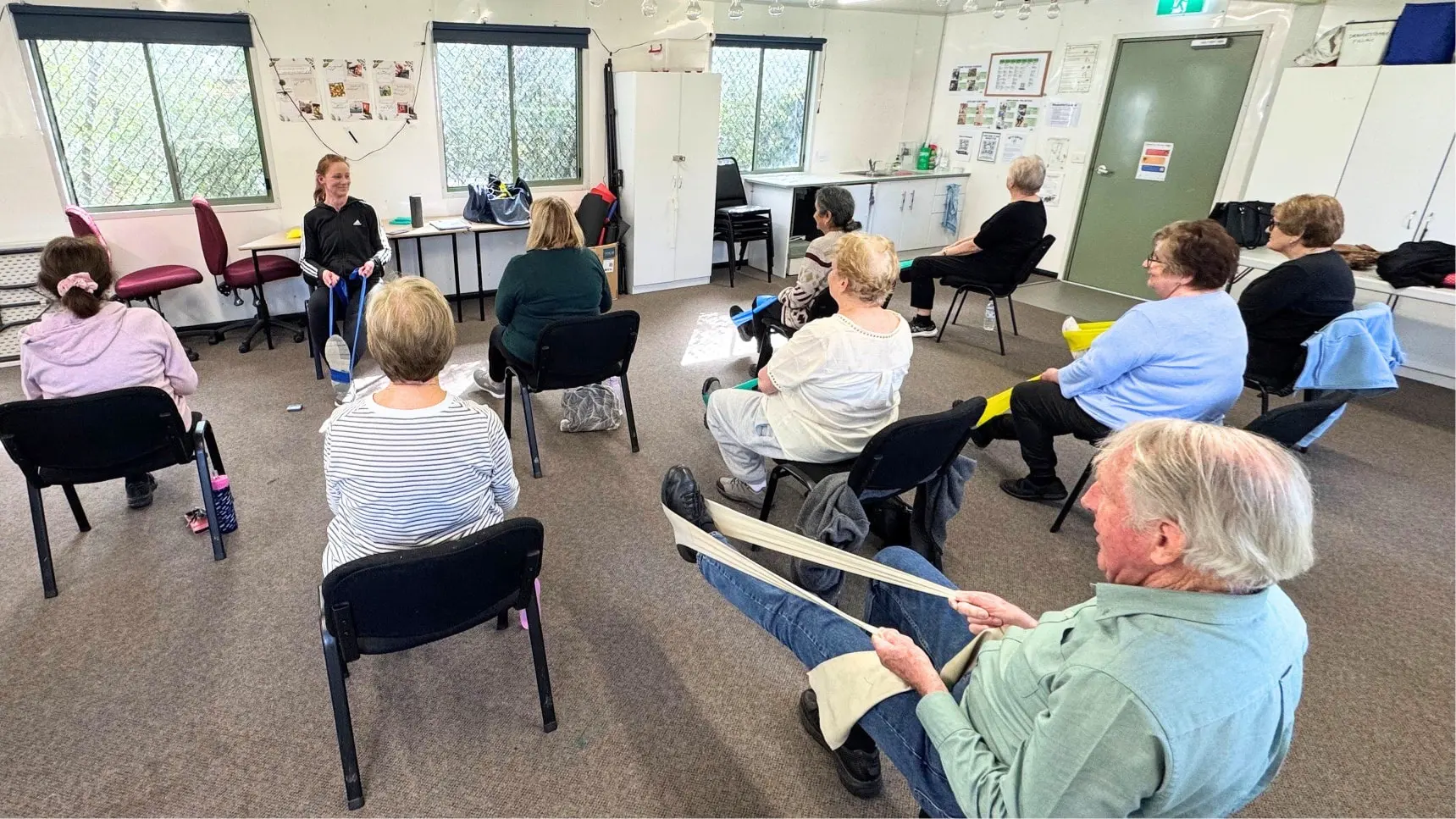 About a dozen people sitting on chairs in a circle in a bright room, stretching resistance bands, with the instructor standing at the front holding one up