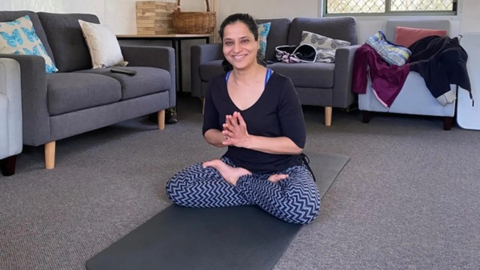 A woman sitting cross-legged on a yoga mat in the lounge room, hands together at her chest, smiling at the camera
