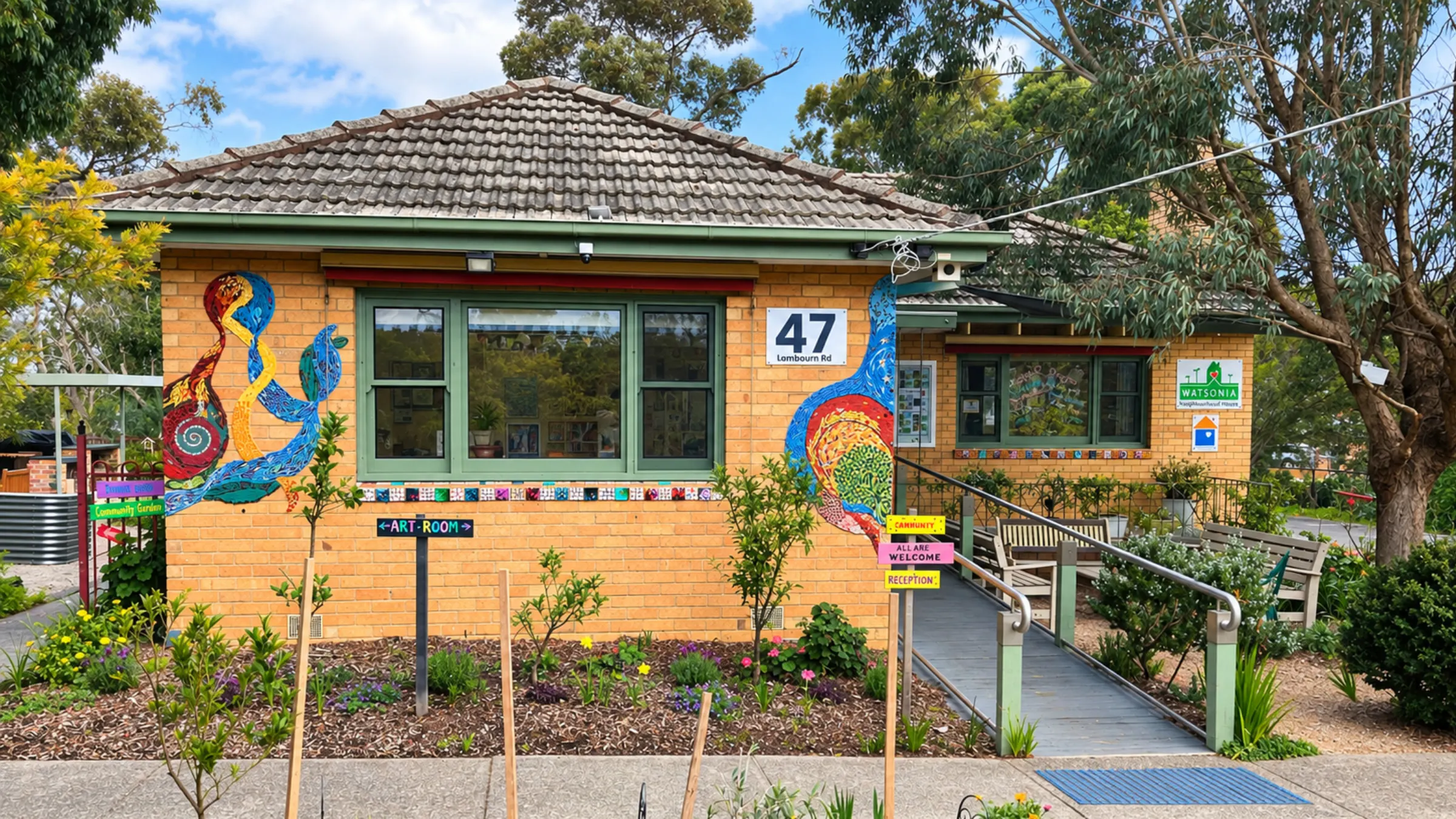 The neighbourhood house from the footpath on a clear day: a brick building under gum trees, with a big mosaic panel either side of the front window, a sign reading 47 Lambourn Rd, a ramp up to the entrance and a garden bed in flower along the path