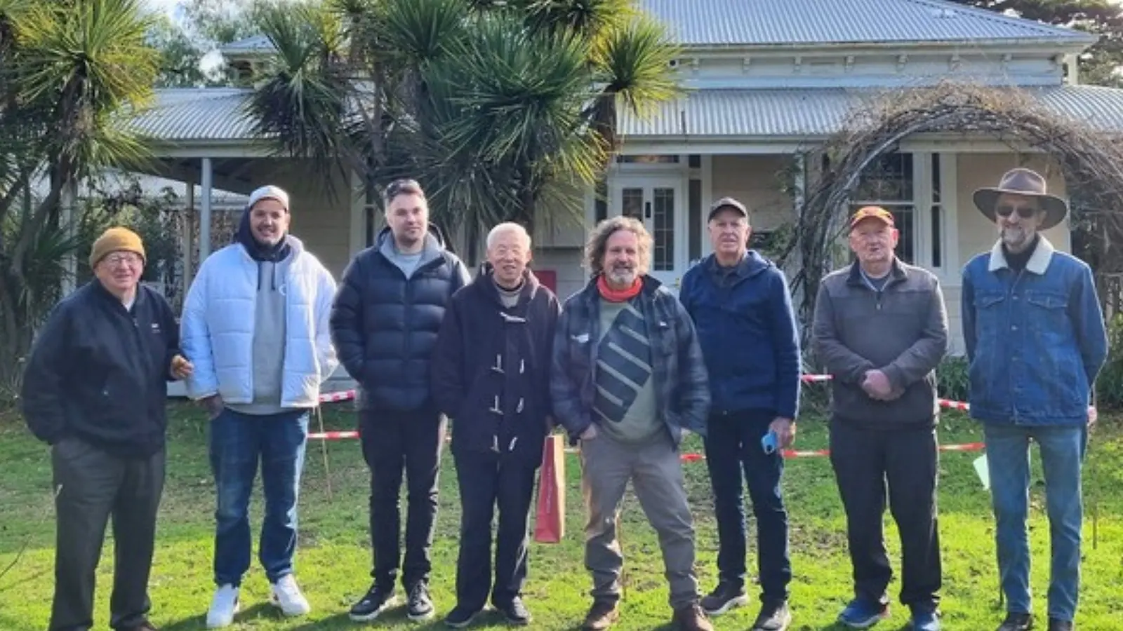 Eight men from the group standing in a row on the grass in front of a weatherboard heritage house, rugged up in jackets and hats