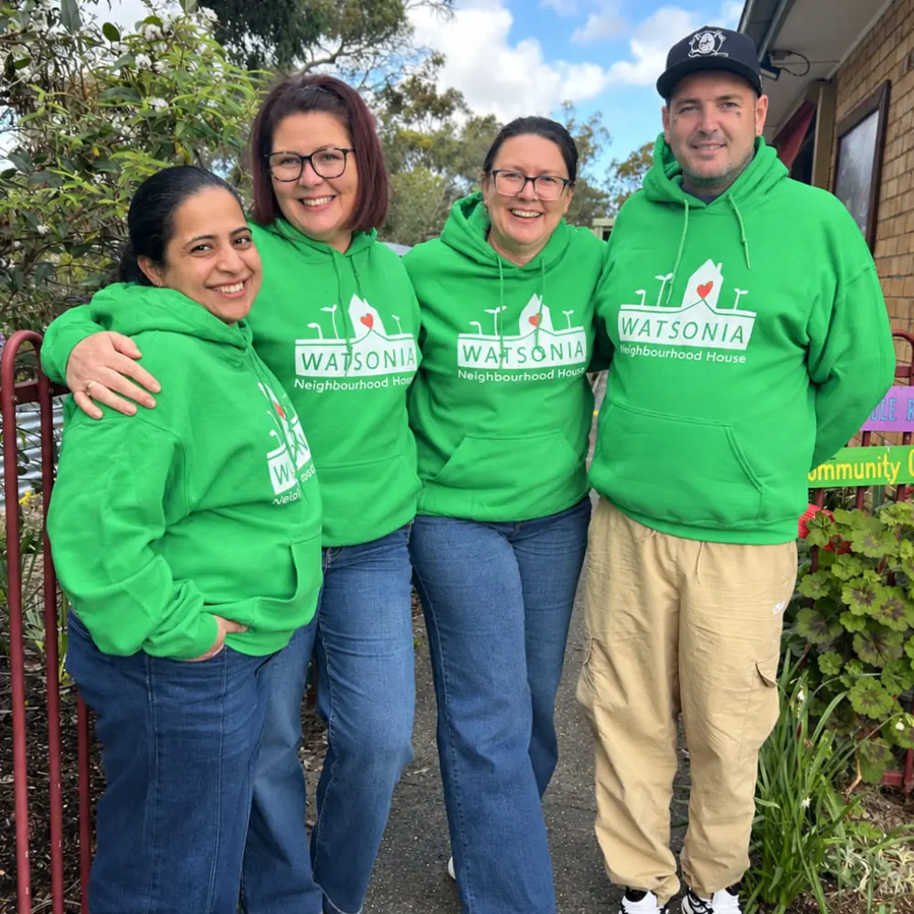 Four people in bright green Watsonia Neighbourhood House hoodies, standing arm in arm by the community garden gate and smiling at the camera