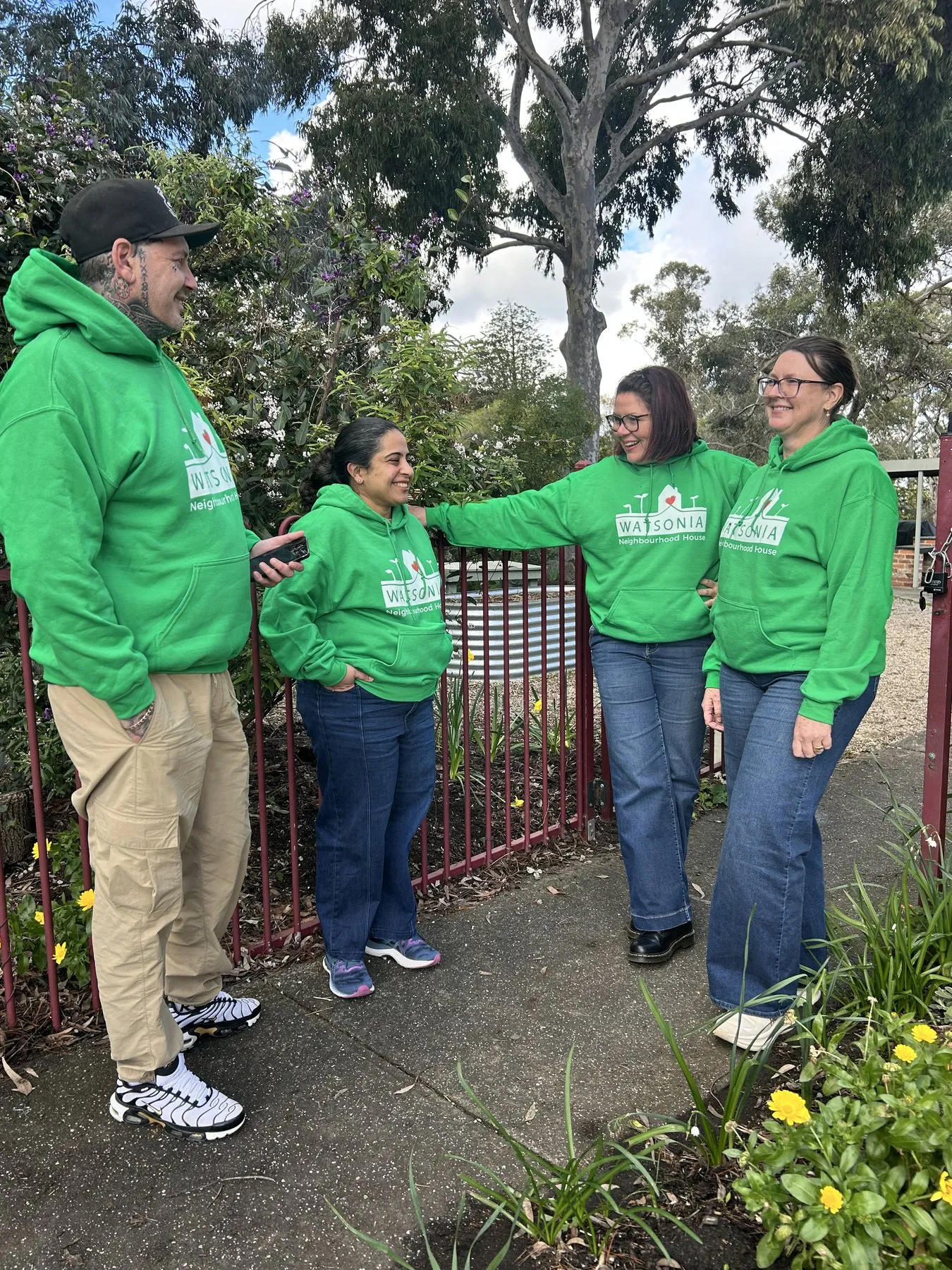 Four staff in green Watsonia Neighbourhood House hoodies laughing together beside the red front gate, daffodils and a large gum tree behind them