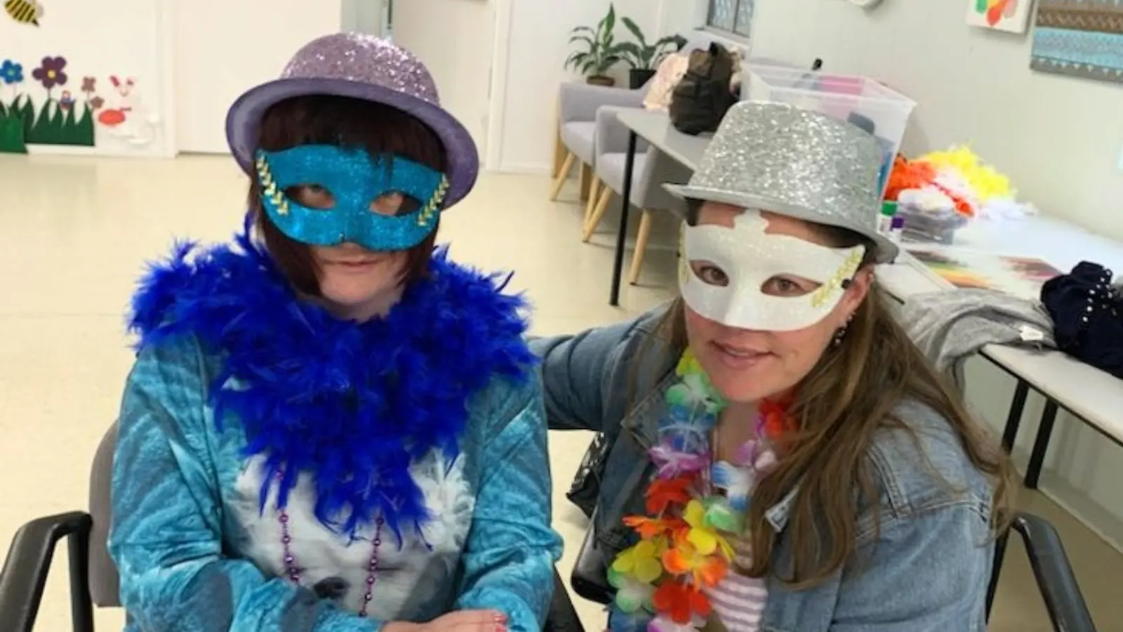 Two women sitting side by side in the activity room, wearing sparkly party hats, glittery eye masks and feather boas