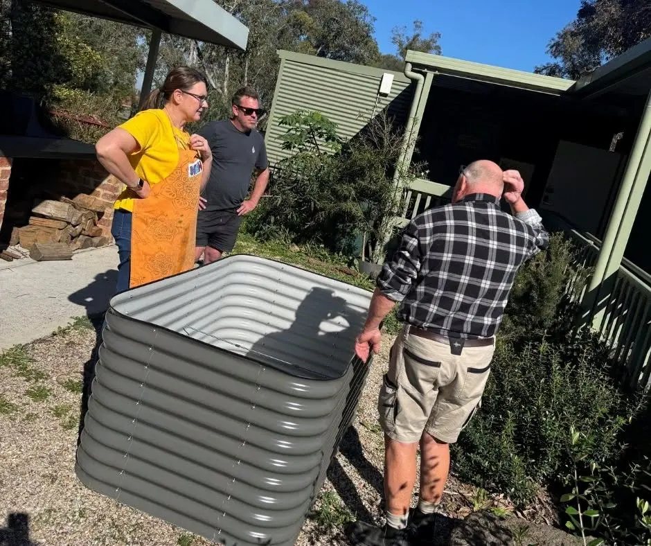 Three people standing around a new corrugated steel raised bed in the garden, beside the covered barbecue area